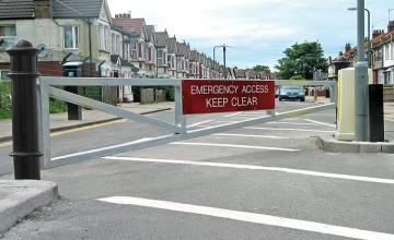 Emergency access gate with Keep Clear sign. A silver gate blocks the road, with houses in the background. Choosing an access gate.