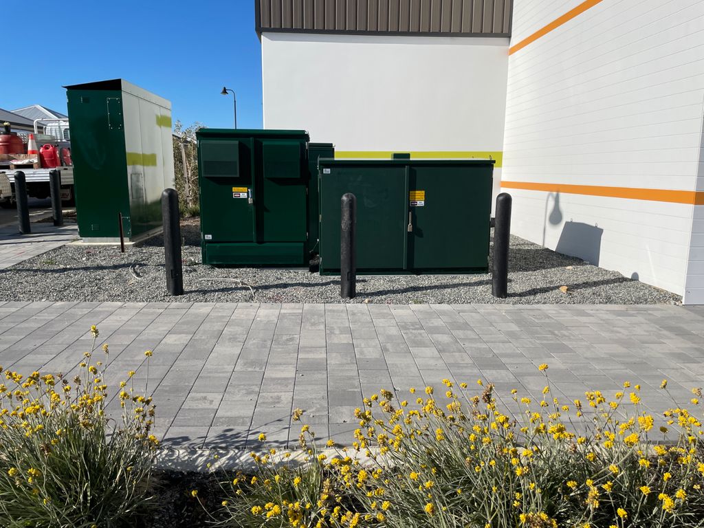 BOR Bollards 1 Non Conductive Bollard 150mm Round 2 Outdoor shot featuring Non Conductive 150mm Round Bollard posts protecting utility boxes near a building, with yellow flowers in the foreground.