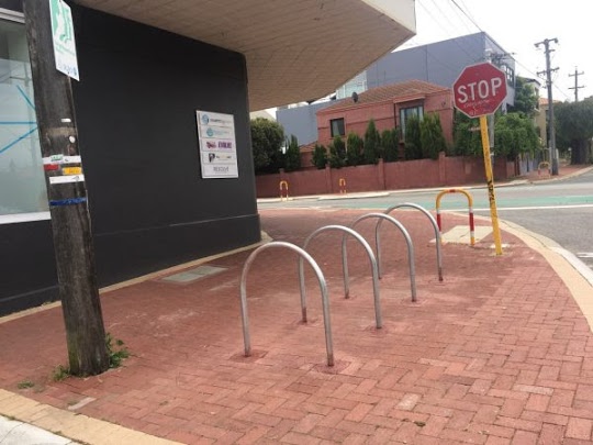 Bike racks outside a business refine its image. Four U-shaped racks on a brick sidewalk near a stop sign. The racks are silver and the sidewalk is red.