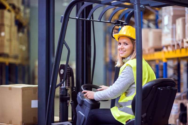 Forklift Safety Cages Essential Considerations For Improved Forklift Safety 1 Forklift safety image: Smiling woman operator in hard hat and safety vest driving a forklift in a warehouse with shelves of boxes.