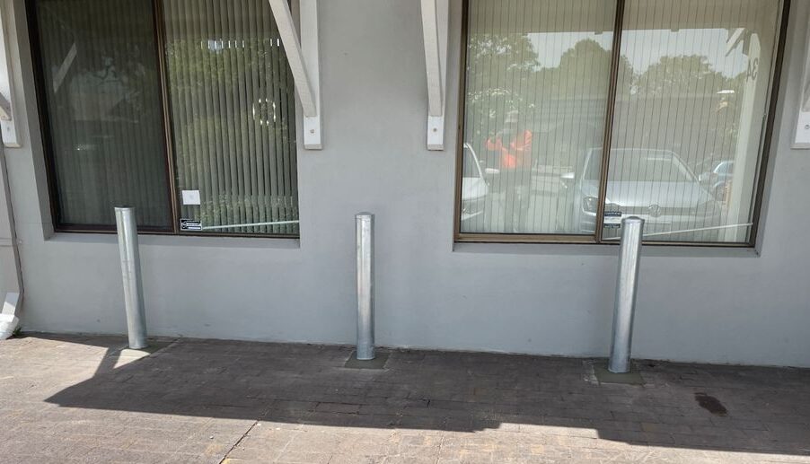 Galvanised Bollards installed e1710290099298 Dugite 114mm x 1500mm In Ground Bollard Galvanised 2 Three Dugite 114mm x 1500mm In Ground Galvanised Bollards stand outside a building with windows, providing security and protection.