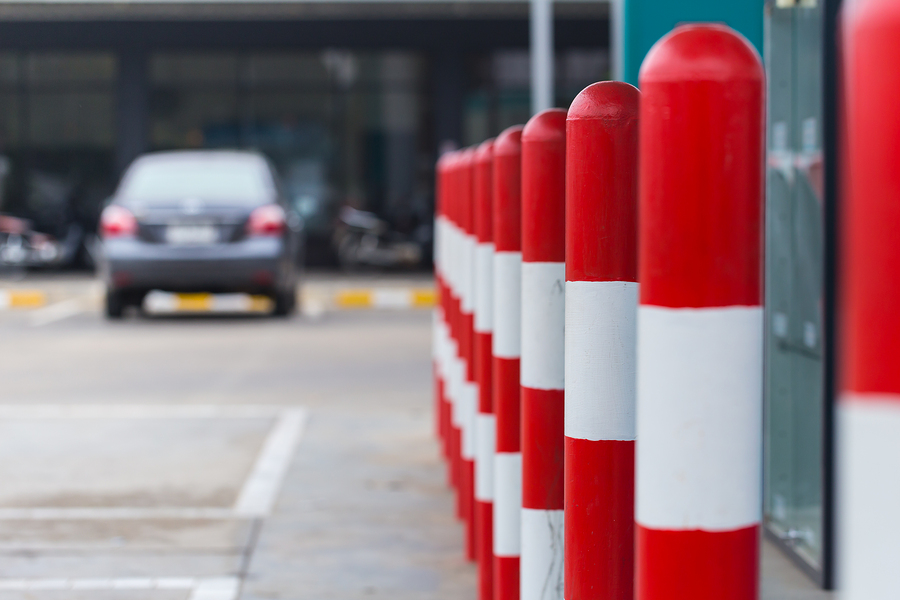 Red and white parking bollards line a car park to improve car park safety. A blurred car is parked in the background, in a parking spot.
