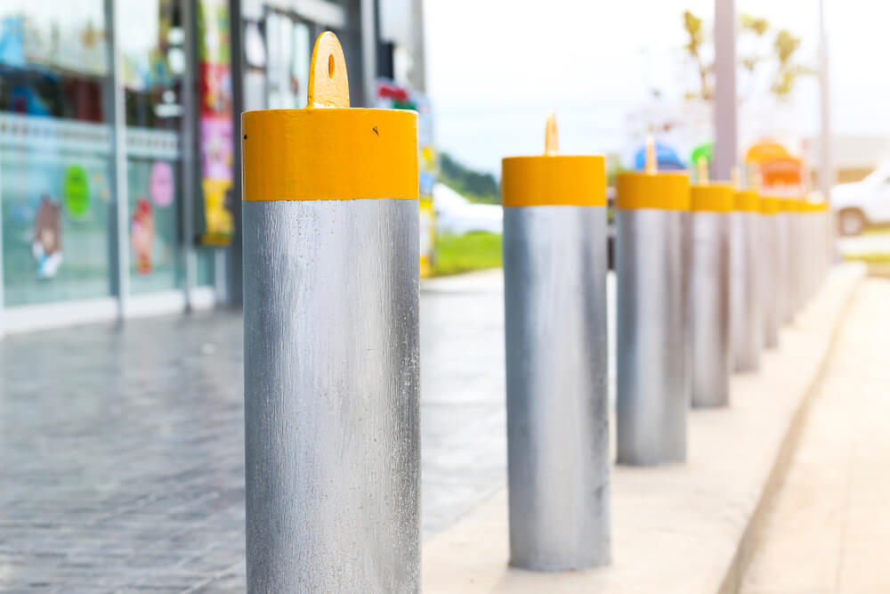 Silver bollard installation with yellow tops line a sidewalk. Bollards are used for safety and security, and affect bollard cost.