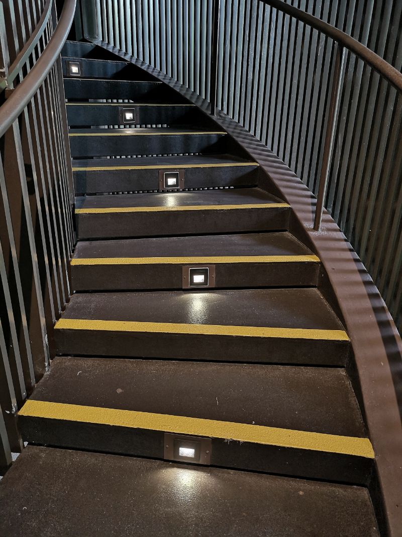 Spiral staircase with anti-slip stair nosing. Yellow safety strips and embedded lights enhance visibility on each step, improving safety.