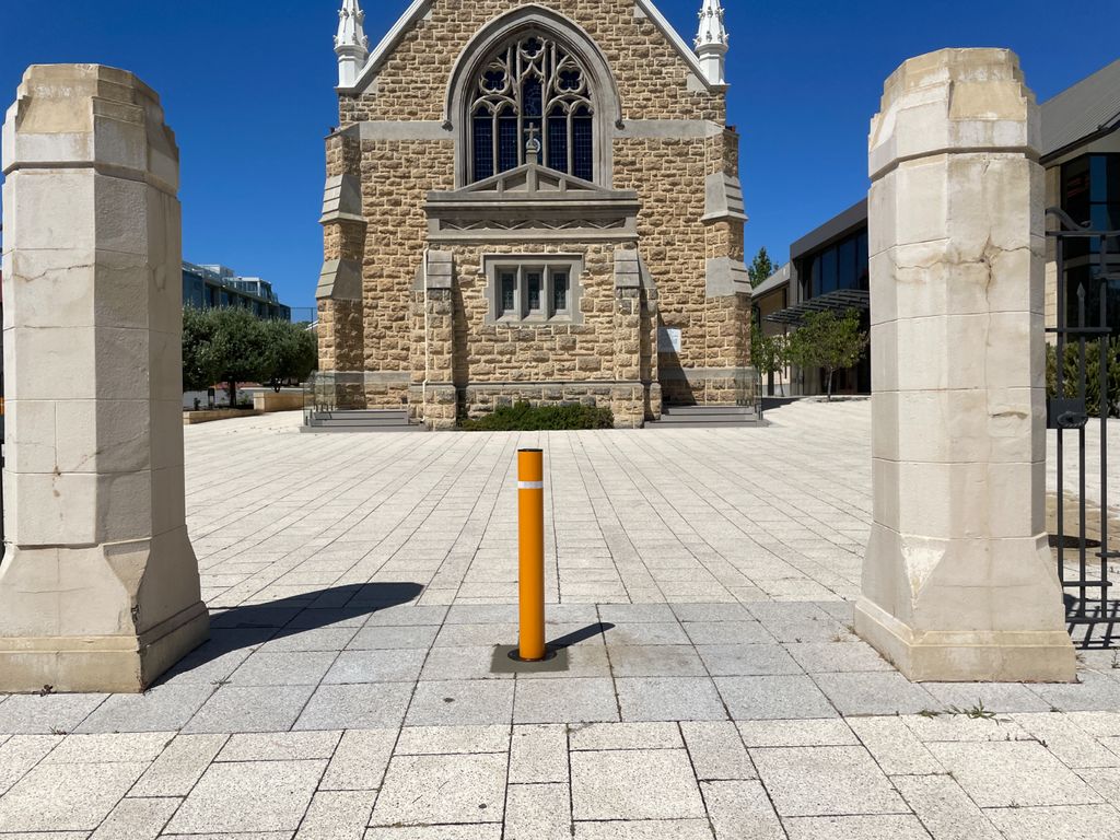 dpr900sy Dugite 114mm x 900mm Retractable Bollard Round Safety Yellow 2 Dugite 114mm x 900mm Retractable Bollard in Safety Yellow. Round bollard between stone pillars, paving stone courtyard, church in background.