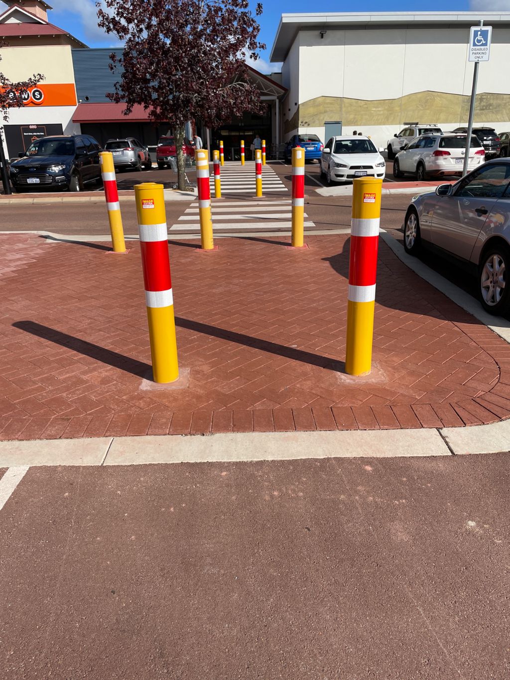 Dugite 165mm x 1800mm In Ground Bollard Safety Yellow MRD, showing safety bollards on a brick crosswalk in front of a building and parked cars.