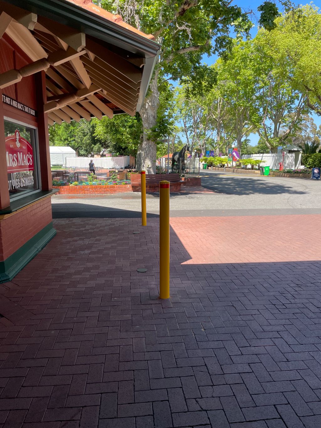 image 6 Dugite 114mm x 1800mm In Ground Bollard Safety Yellow 2 Safety Yellow Dugite 114mm In Ground Bollard. Outdoor view of yellow bollards on a brick pathway, trees and buildings in the background.