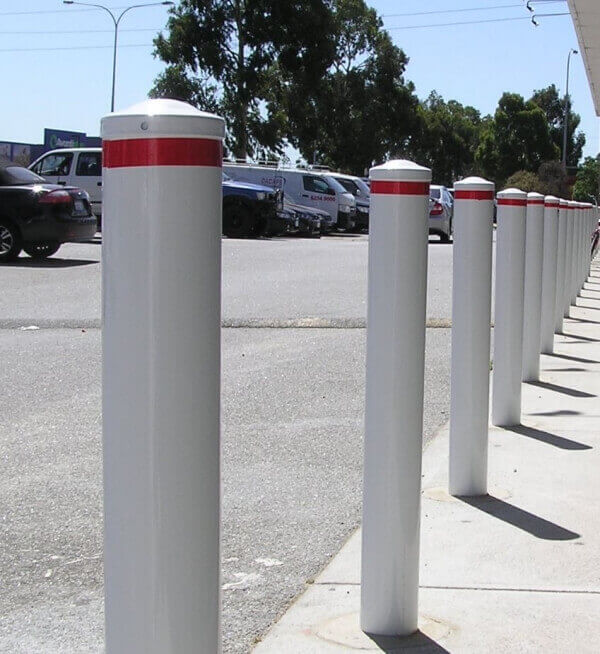 A row of white safety bollards with red stripes, used for traffic and pedestrian safety. Parking lot with cars and trees in the background.