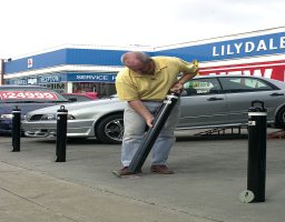 scaled 500d12f7e092a How to Place Removable Bollards 1 Man installing removable bollards on a concrete surface. Black bollards are placed in a line, with cars and a building in the background.