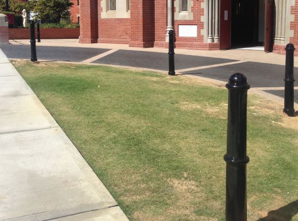 Black cast iron bollards line a walkway and lawn in front of a brick building. These sustainable bollards protect pedestrians and property.