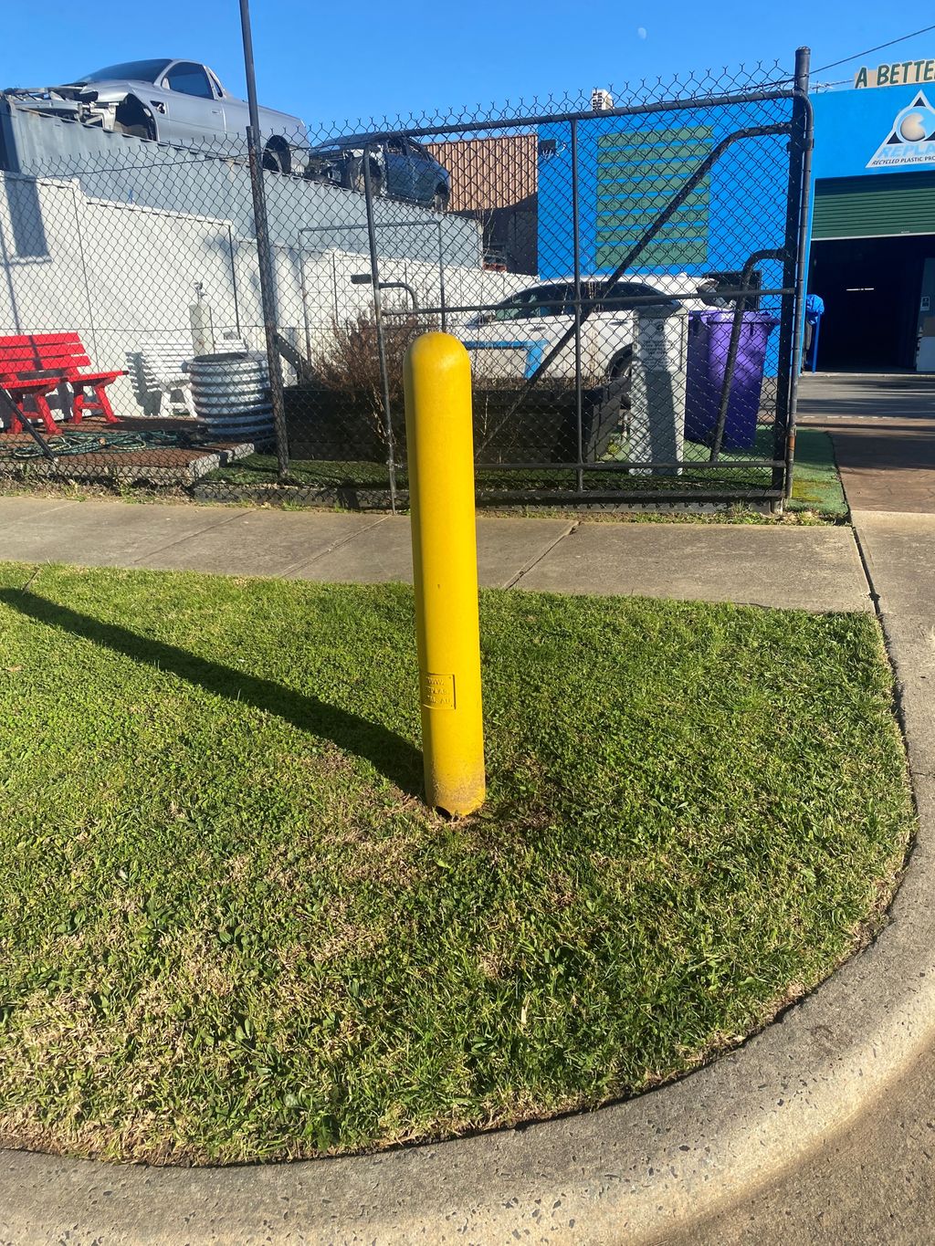 BOR150 Yellow Non Conductive Bollard 150mm Round - Coloured Blue or Yellow 3 Yellow non conductive bollard, 150mm round, stands in green grass. A fence, cars, and a building are in the background on a sunny day.